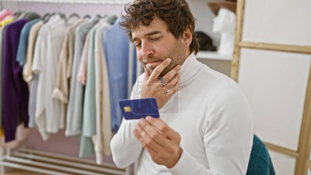 Photo for A contemplative young man with a beard examines a credit card in a modern clothing room, surrounded by stylish garments. - Royalty Free Image