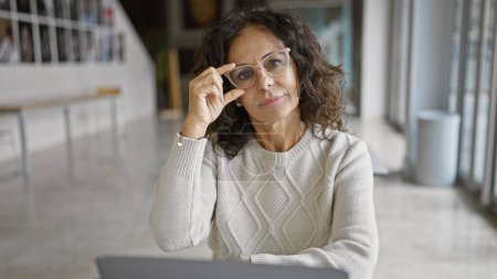 Téléchargez les photos : Femme hispanique mature avec des lunettes dans un cadre de bureau en regardant la caméra, exsudant la confiance professionnelle. - en image libre de droit