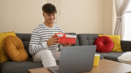 Foto de Sorprendido joven hispano sentado en casa en el sofá, desempacando el regalo sorpresa mientras hablaba felizmente y sonreía durante la videollamada en línea. - Imagen libre de derechos