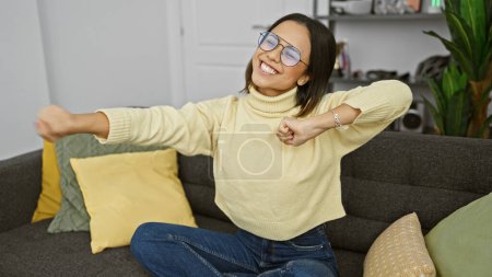 Photo for Happy young hispanic woman stretching on a cozy couch in a modern living room, wearing glasses and casual clothing. - Royalty Free Image
