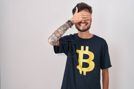 Photo for Young hispanic man with tattoos wearing bitcoin t shirt smiling and laughing with hand on face covering eyes for surprise. blind concept. - Royalty Free Image