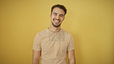 Téléchargez les photos : Beau jeune homme hispanique souriant sur un fond jaune, respirant confiance décontractée et charme. - en image libre de droit