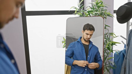 Photo for Handsome hispanic man buttoning shirt in a modern dressing room with plants and hats. - Royalty Free Image