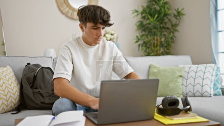 Estudiante joven hispano concentrado, absorto en la educación en línea, sentado en el acogedor sofá de la sala de estar, centrado en la pantalla del ordenador portátil, relajarse en casa, rodeado de tecnología