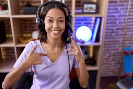 Téléchargez les photos : Jeune femme hispanique jouant à des jeux vidéo portant un casque pointant du doigt un sourire heureux et fier - en image libre de droit