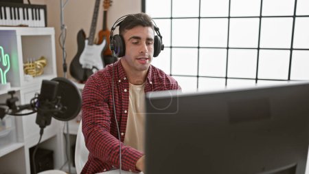 Photo for Handsome young hispanic man, a professional musician immersed in melody, sitting relaxed in the interior of a music studio wearing headphones, using a computer for his acoustic performance - Royalty Free Image