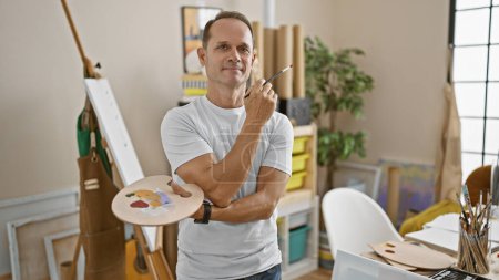 Photo for Confident middle-aged hispanic artist, reveling in creativity, strikes a relaxed pose with palette and paintbrush in hand amidst the warm ambiance of his college art studio - Royalty Free Image
