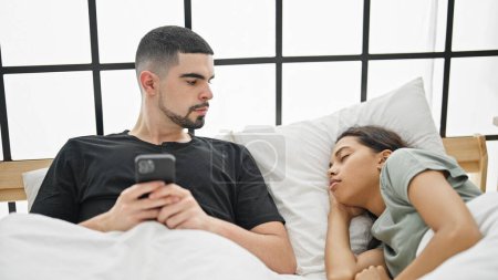 Foto de Encantadora escena de hermosa pareja relajándose cómodamente en la cama, hombre mensajes de texto en el teléfono inteligente como su novia duerme en su dormitorio decorado con amor - Imagen libre de derechos
