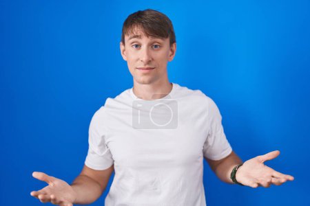 Foto de Hombre rubio caucásico de pie sobre fondo azul despistado y confundido con los brazos abiertos, sin concepto de idea. - Imagen libre de derechos