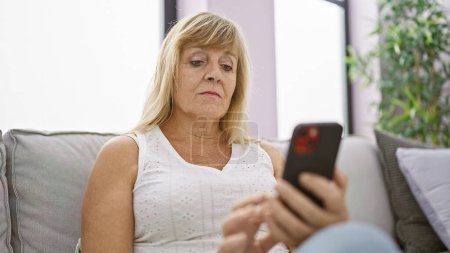 Ernste blonde Frau mittleren Alters kämpft mit der Technik, verärgerter Gesichtsausdruck, während sie drinnen auf dem Sofa sitzt, Nachrichten auf dem Smartphone tippt und in das Problem vertieft ist, das in ihrem stilvollen Wohnzimmer vor sich geht.