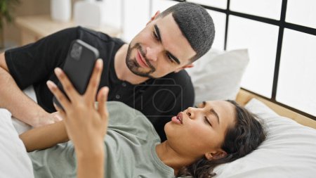 Photo for Relaxed beautiful couple, lying in bed, smiling and texting love messages on their smartphone in cozy bedroom - Royalty Free Image