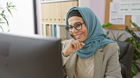 Téléchargez les photos : Femme professionnelle souriante avec hijab en utilisant l'ordinateur dans un cadre de bureau moderne - en image libre de droit