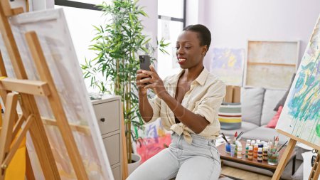 Téléchargez les photos : Artiste afro-américaine confiante, dessinant à partir d'une image réalisée avec son smartphone dans un studio d'art confortable - en image libre de droit