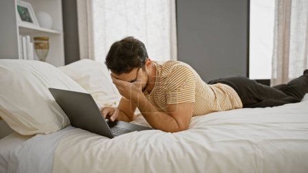 Photo for A stressed hispanic man lying on a bed using a laptop in a modern bedroom setting, exuding a sense of frustration and exhaustion. - Royalty Free Image