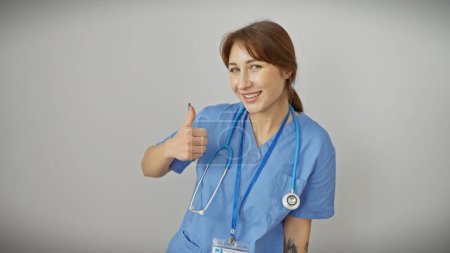 Photo for Confident young caucasian female nurse in blue scrubs giving thumbs up against a white isolated background - Royalty Free Image