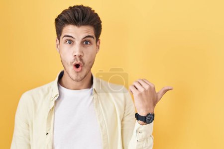 Photo for Young hispanic man standing over yellow background surprised pointing with hand finger to the side, open mouth amazed expression. - Royalty Free Image