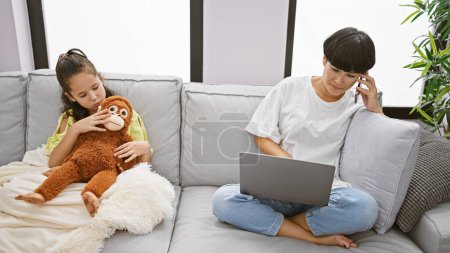 Photo for Mother-daughter bonding at home, woman working while lovely girl sits on sofa hugging monkey doll, embracing casual lifestyle - Royalty Free Image