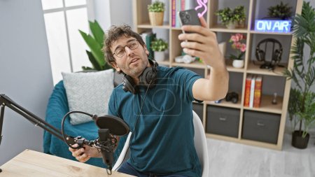 Téléchargez les photos : Un homme hispanique prend un selfie dans un studio de radio avec un équipement de podcasting moderne et une pancarte "en ondes". - en image libre de droit