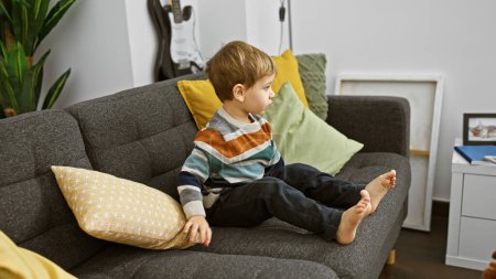 Téléchargez les photos : Bébé blond assis calmement sur un canapé gris dans un salon moderne, regardant pensivement. - en image libre de droit