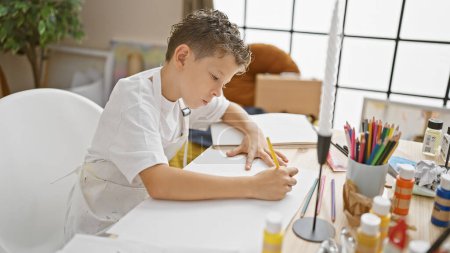 Téléchargez les photos : Adorable petit garçon artiste blond dessinant intensément dans son carnet, immergé dans la classe d'art au studio, avec pinceaux, toile et palette autour de lui. - en image libre de droit