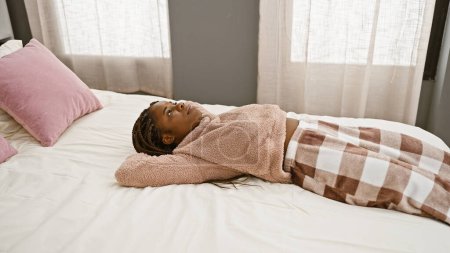 Eine ruhige afrikanische Frau mit Zöpfen liegt besinnlich in einem gemütlichen Schlafzimmer, eingewickelt in eine Decke auf einem Bett.