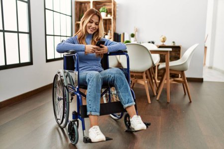 Téléchargez les photos : Jeune femme caucasienne utilisant smartphone assis sur fauteuil roulant à la maison - en image libre de droit