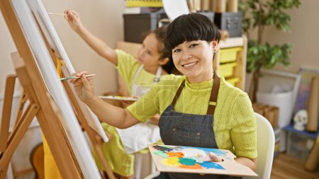 Profesora de arte confiada y estudiante pintando alegremente juntos, sonrisas floreciendo en el acogedor interior del estudio de arte.