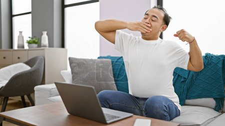 Photo for Exhausted young chinese man sitting on a sofa, yawning while using his laptop at home - Royalty Free Image