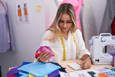 Foto de Young blonde woman tailor writing on notebook holding cloth at clothing shop - Imagen libre de derechos