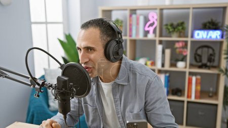 Handsome hispanic man speaking into microphone in a radio studio setting, wearing headphones.