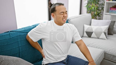 Besorgter junger Chinese, der unter schweren Rückenschmerzen leidet, zu Hause unglücklich auf dem Sofa sitzt und die harte Realität von Wirbelsäulenverletzungen zeigt.