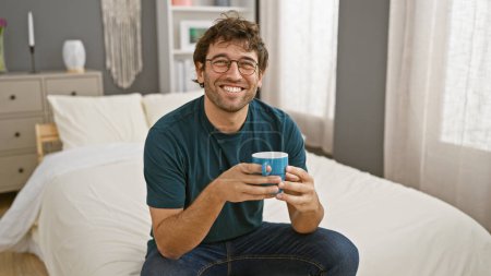 Foto de Sonriente joven hispano con barba sosteniendo una taza en un acogedor dormitorio. - Imagen libre de derechos