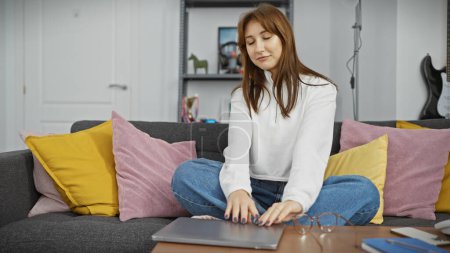 Photo for A young woman relaxes on a couch with colorful cushions, using a laptop in a cozy modern living room. - Royalty Free Image