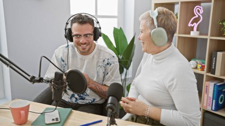 Photo for A young man and an elderly woman wearing headphones laugh together during a podcast recording in a modern studio. - Royalty Free Image