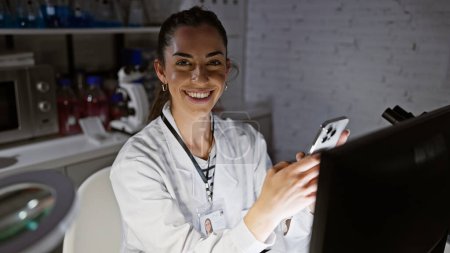 Téléchargez les photos : Jeune belle scientifique hispanique utilisant ordinateur et smartphone souriant au laboratoire - en image libre de droit