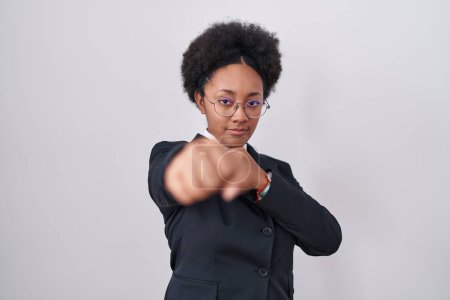 Téléchargez les photos : Belle femme africaine aux cheveux bouclés portant une veste d'affaires et des lunettes poinçon poing pour se battre, attaque agressive et en colère, menace et violence - en image libre de droit