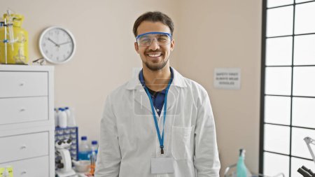 Photo for Smiling hispanic man wearing lab coat and safety goggles in a bright laboratory setting - Royalty Free Image