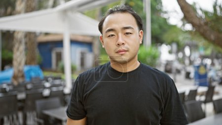 Photo for Cool, handsome asian guy standing on a sunny coffee shop terrace in the heart of the city, showing a serious expression while shooting a relaxed urban lifestyle. - Royalty Free Image