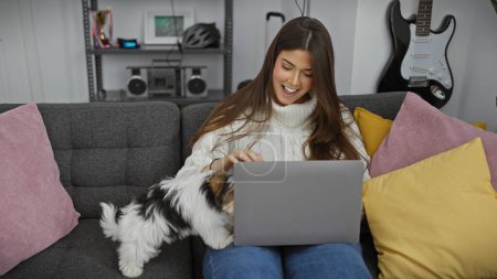 Photo for Smiling hispanic woman using laptop with biewer terrier on couch in modern living room. - Royalty Free Image