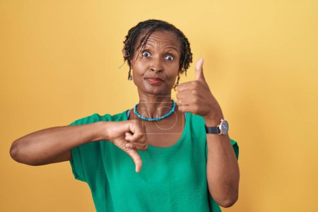 Foto de Mujer africana con rastas de pie sobre fondo amarillo haciendo pulgares arriba y abajo, desacuerdo y expresión de acuerdo. conflicto loco - Imagen libre de derechos