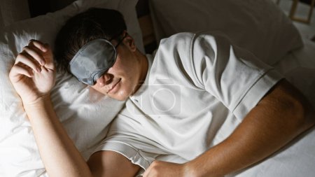 Téléchargez les photos : Nuit confortable, jeune adolescent hispanique épuisé jouissant d'un sommeil reposant, détendu et confortable au lit avec masque de sommeil, dans une chambre chaude et faiblement éclairée - en image libre de droit