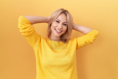 Foto de Mujer caucásica joven usando suéter amarillo relajante y estiramiento, brazos y manos detrás de la cabeza y el cuello sonriendo feliz - Imagen libre de derechos