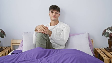 Photo for Handsome young hispanic man in pyjamas, awake, comfortably resting on his cozy bed, displaying an intense, serious expression, deep in thought in his relaxed indoor apartment bedroom. - Royalty Free Image