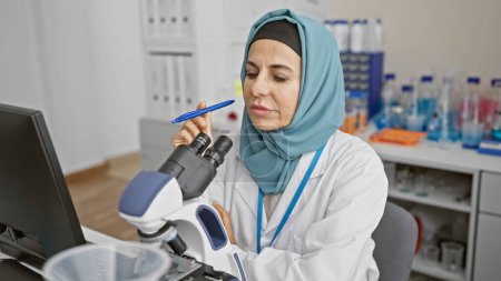 Photo for A woman scientist in a hijab examines a slide under the microscope at a laboratory. - Royalty Free Image