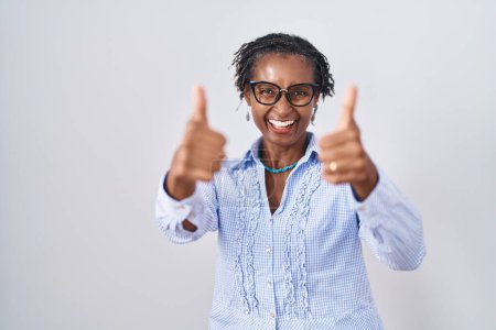 Foto de Mujer africana con rastas de pie sobre fondo blanco con gafas que aprueban hacer gesto positivo con la mano, pulgares arriba sonriendo y feliz por el éxito. gesto ganador. - Imagen libre de derechos