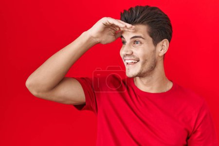 Photo for Young hispanic man standing over red background very happy and smiling looking far away with hand over head. searching concept. - Royalty Free Image
