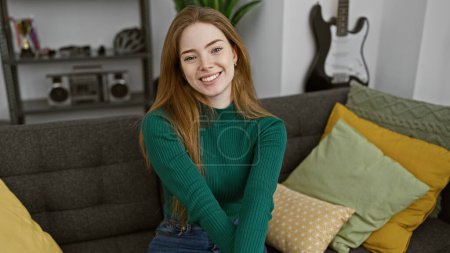 A smiling young blonde woman dressed in a green sweater sits comfortably indoors on a gray couch in a modern living room.