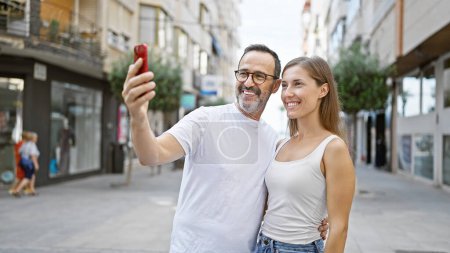 Père et fille confiants partagent un moment agréable, prenant joyeusement un selfie cool sur leur téléphone portable debout dans une rue animée de la ville.