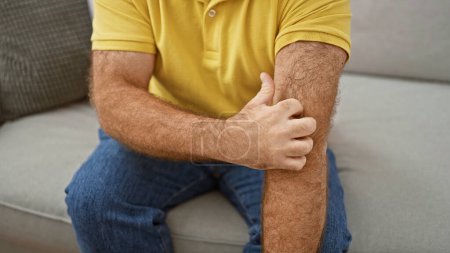 Photo for Handsome middle-aged caucasian man sitting on sofa at home, scratching irritated skin on arm. itchy dermatitis rash causing discomfort. health care needed for allergic reaction. - Royalty Free Image