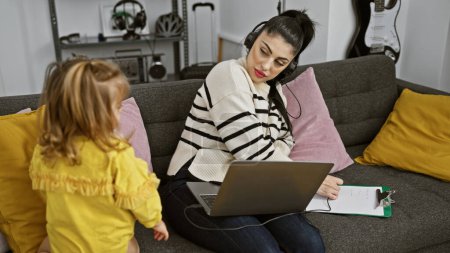 Foto de Una mujer trabaja en el portátil con auriculares en una sala de estar moderna, mientras que su hija en un vestido amarillo está al lado del sofá. - Imagen libre de derechos